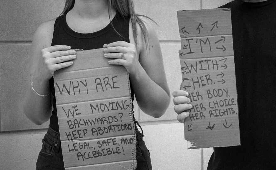 A couple stands together holding coordinating signs. The husband emphasizes that it is his wife’s choice what she does with her body.