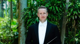 A man in a tuxedo stands in the midst of green trees.