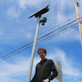 Jack Rankin, under a Flock camera in a parking lot outside a cafe in Eastport. The camera takes surveillance pictures of people driving east on M-88. (Photo: Austin Rowlader / IPR News)
