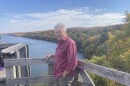 A man in a red shirt and cargo pants stands leaning against a fence. Behind him is a river and line of trees.