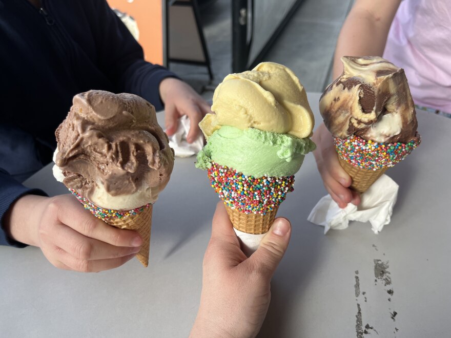 Three hands hold waffle cones filled with scoops of ice cream in assorted flavors, each cone dipped in rainbow sprinkles, over an outdoor café table.