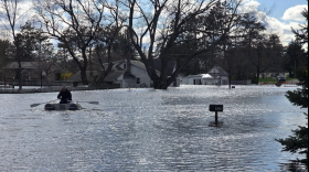 Man paddles on a boat through flood waters in Cadillac, Michigan. Behind him are two white houses submerged in water. 