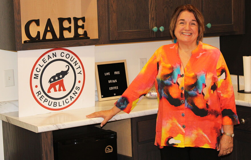 Woman in multi-colored top standing with her right hand placed on a countertop next to a sign that reads 'McLean County Republicans' and shows a logo of an elephant.