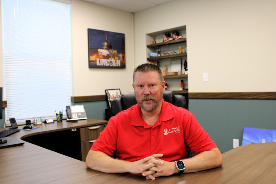 A man with short hair and a beard sits at a desk in an office, wearing a red collared shirt and smartwatch. Shelves, a desk phone, and a Lincoln artwork are visible in the background.