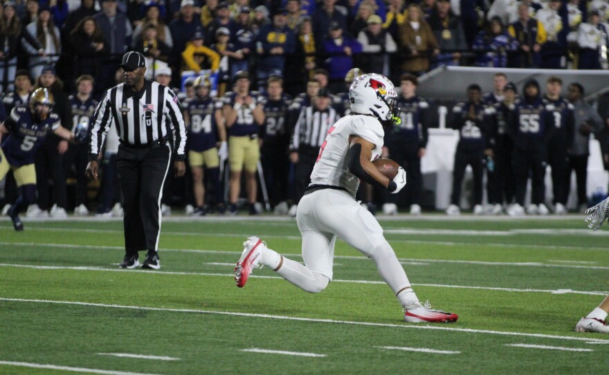 A football player in a white uniform runs with the ball during a game, with a referee and opposing players in dark uniforms visible in the background, and a crowd watching from the stands.