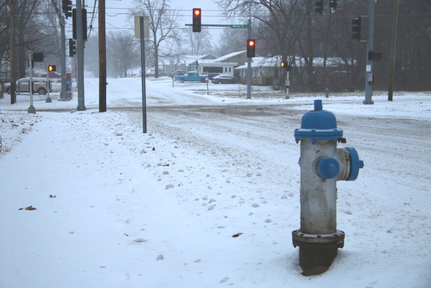 A blue and silver fire hydrant is in the right side of the frame, snow surrounds it. In the background are traffic lights and an intersection. The roads are snow covered.