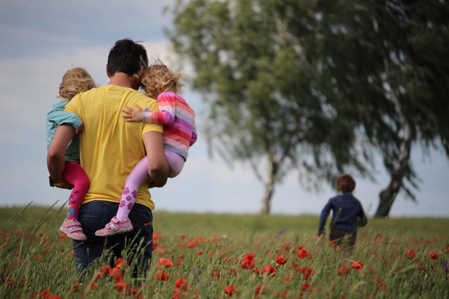 A man in a field, facing away from the camera and holding two children, as another child runs away from the camera.