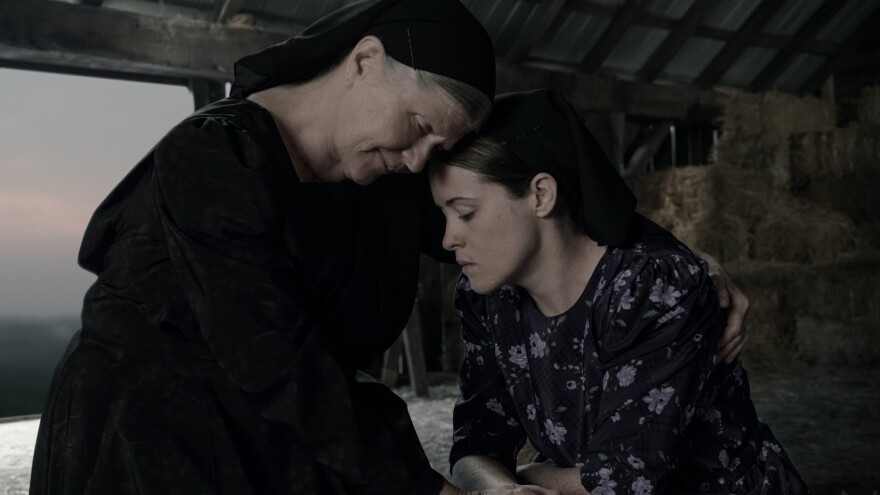 An older woman and a younger woman, both in modest clothing, embrace with their eyes closed while sitting in a barn with hay bales in the background.