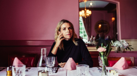 Aoife O'Donovan sits at a dining table in a pink room, looking at the camera with one hand on her cheek.
