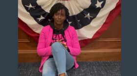 Black woman sitting on the floor, leaning against a wood-paneled desk draped with red, white and blue bunting. She wears a bright pink blazer.