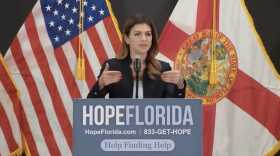 Woman with long brown hair standing behind a podium that says Hope Florida with U.S. and Florida flags behind her