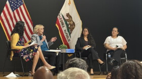 Four people sitting in chairs. The second person from the left is holding a microphone. Behind them are two flags.