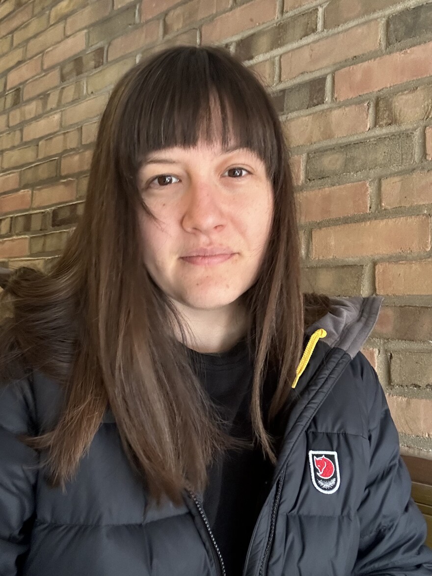 Woman with brown hair passed her shoulders and bangs wearing a fluffy black coat with a backdrop of a brown and beige brick wall.