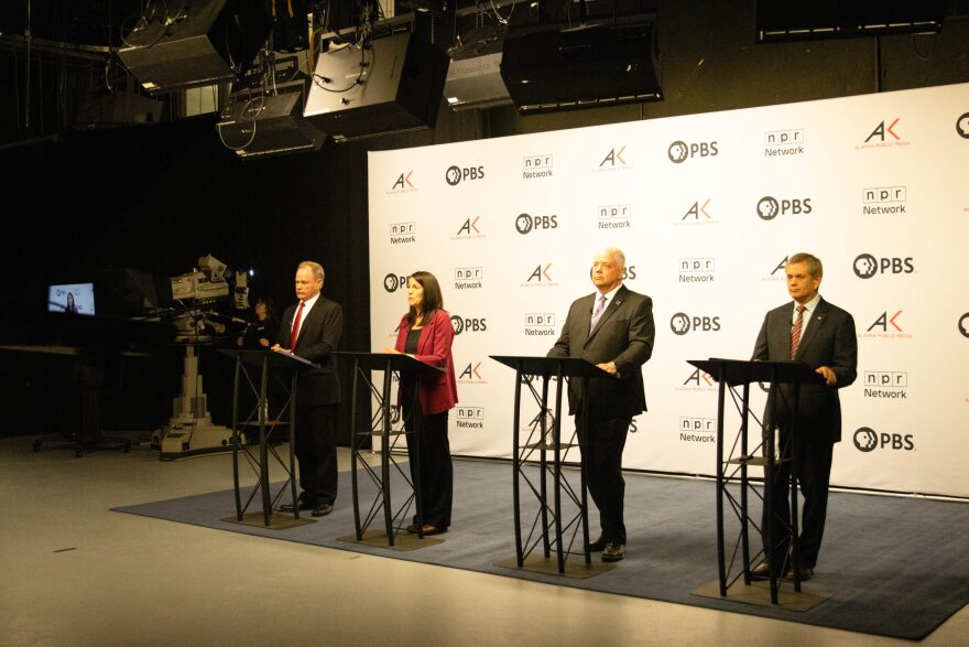 Four people, a man in a suit with red tie, a woman with a deep red blazer, a man in a suit with purple tie, and a man in a suit with a striped tie stand behind podiums in front of a white backdrop with the logos for PBS, NPR, and Alaska Public Media preparing for a debate.