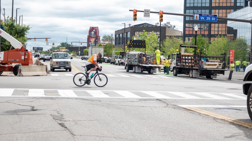 A cyclist wearing reflective gear and a helmet crossing through the Lorain Avenue and West 25th Street intersection surrounded by other vehicles.
