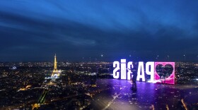 A visitor strolls past a "Paris" sign that is reflected in the glass wall of the Montparnasse tower observation deck as the Eiffel Tower stands in the background ahead of the 2024 Summer Olympics, Tuesday, July 23, 2024, in Paris. (AP Photo/David Goldman)