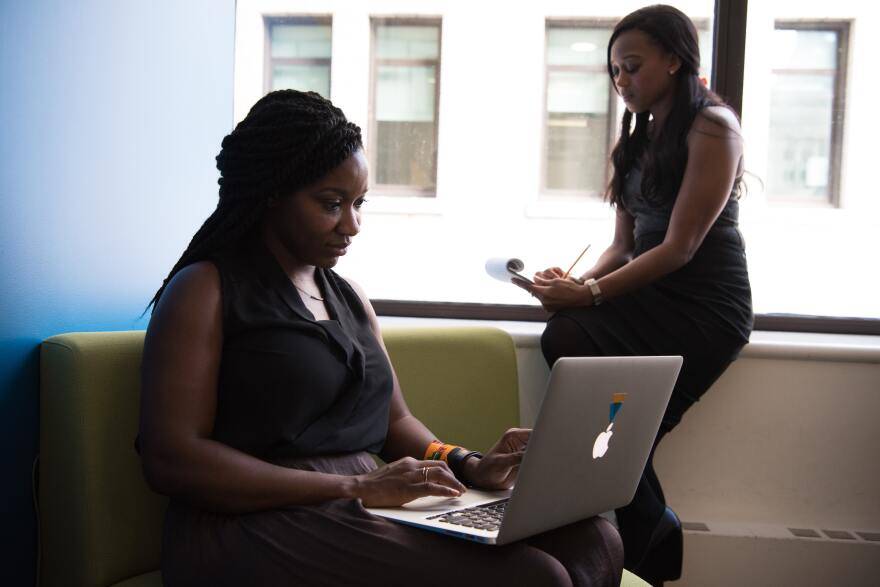 Young Black professional woman in the background sits on the windowsill in an office dressed in black sleeveless top and pants writing on a pad of paper while another Black woman in the foreground sits on a green upholstered chair wearing a sleeveless black dress typing on a laptop.