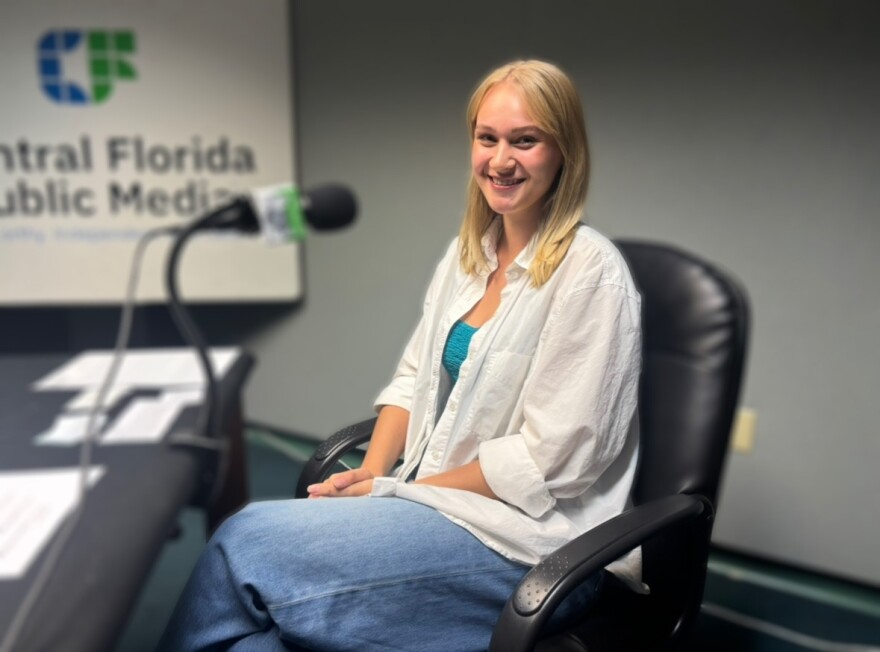 A young woman with blonde hair sits in a chair and smiles, she's sitting in front of a microphone.
