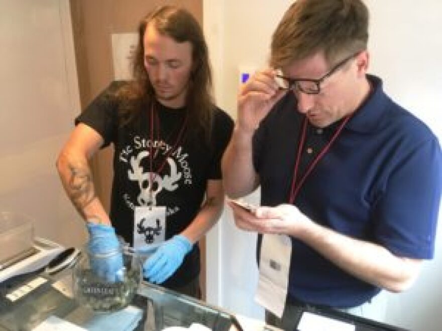 Travis Carter gets some cannabis buds out to measure for a customer while Mark Woodward checks an ID on the opening day for Ketchikan’s Stoney Moose. (KRBD photo by Leila Kheiry)