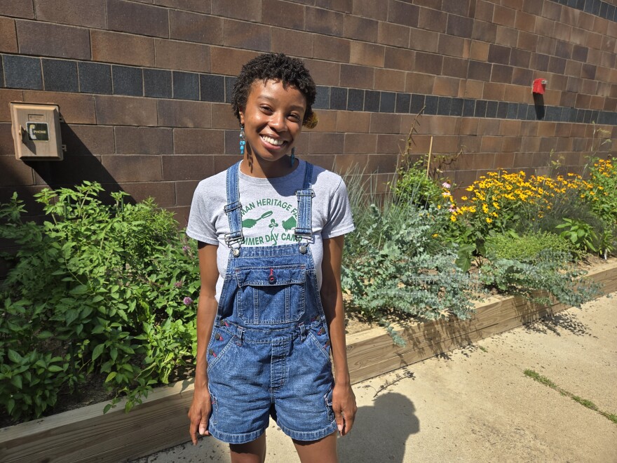 a woman in a gray t-shirt and overalls stands smiling in a garden