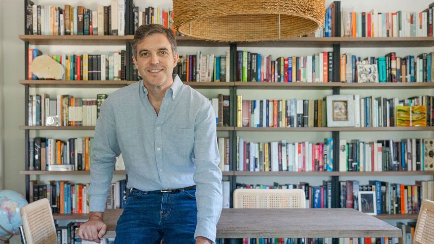 Man in blue jeans sits on edge of dining room table, smiles into the camera with books around him 