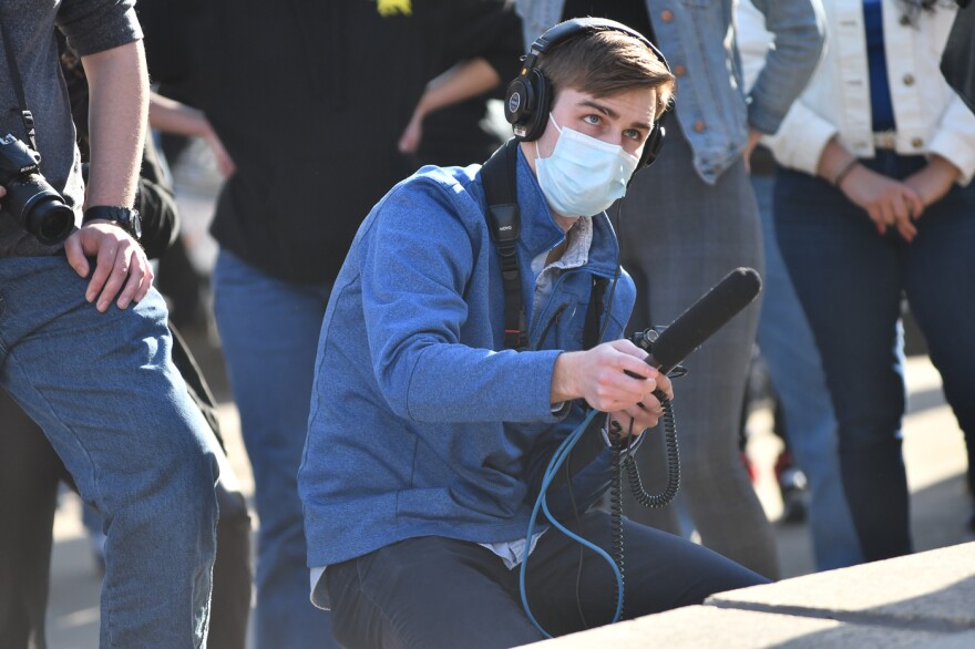 Young white male reporter in long-sleeved denim blue shirt and navy pants wearing a disposable mask is crouched down in front of others who are standing.  He is wearing headphones with his right hand outstretched holding a microphone.