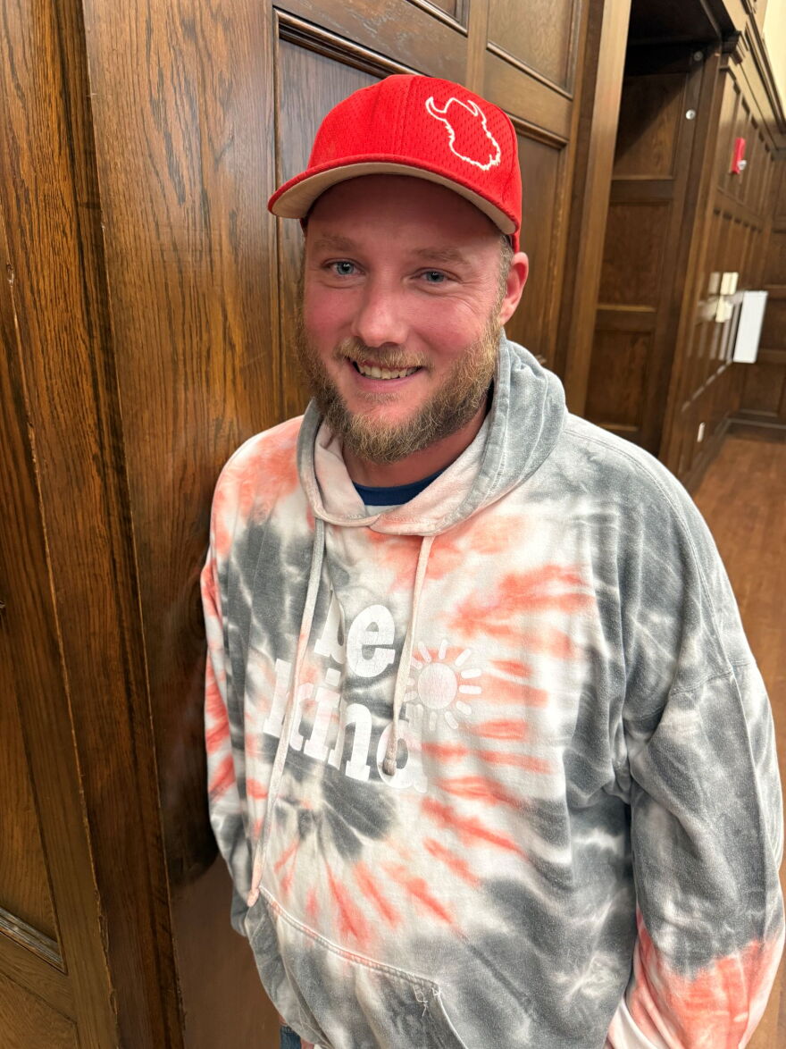 A man in a gray, white and orange tie-dye sweatshirt and a red baseball cap smiles for the camera.