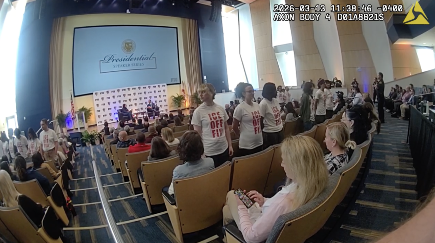 A screenshot taken from police body cam shows several students standing facing the audience with white shirts that say ICE OFF FIU. Two speakers are on stage in the background.