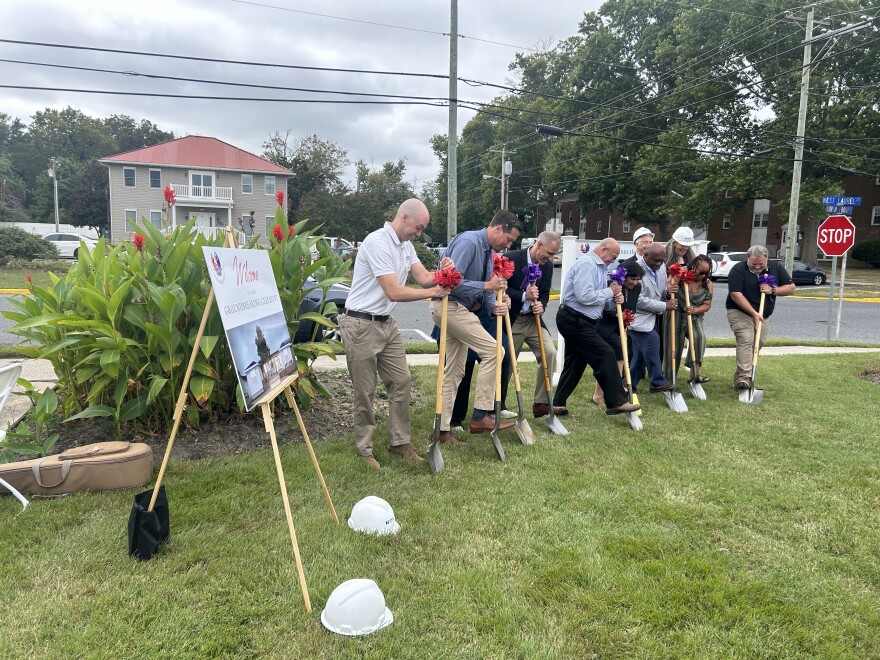 10 people in a line dig shovels into the grass.