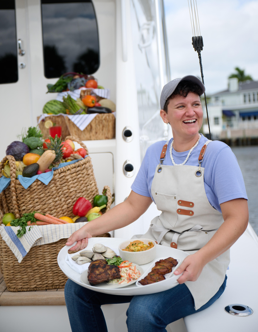 A private yacht chef smiling while serving a platter of gourmet dishes on a yacht, with a basket of fresh vegetables and fruits in the background.