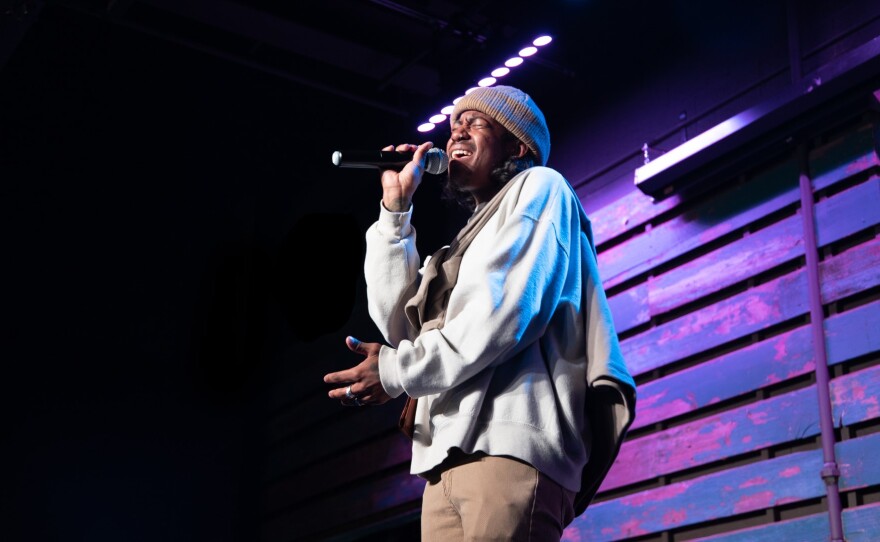 A man sings into a handheld microphone on stage with lighting behind him.