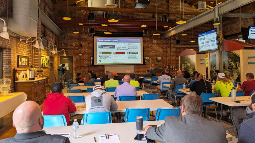 Water utility workers sit at long tables lined up in rows inside a century-old, brick-walled water-pumping station that has been converted into an event space. They look at a large screen and a person speaking at a podium.