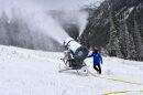 A large snowgun shoots snow on the side of a hill with pine trees in the background.