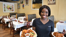 A woman standing inside a small restaurant dining room holds two plates of food. A chicken dish with vegetables is in one hand and gizzards in a red sauce is on the other dish.