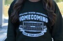 A dark green sweatshirt with white lettering reads "William Woods University Homecoming, Leave Your Legacy" on either side of a printed image of a football field. 