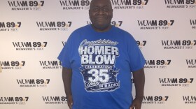 Homer Blow, a long-time radio host in Milwaukee, stands in front of the WUWM 89.7 Milwaukee's NPR photo backdrop wearing a royal blue shirt with the words: "Congratulations Homer Blow Celebrating 35 Years in Radio!"