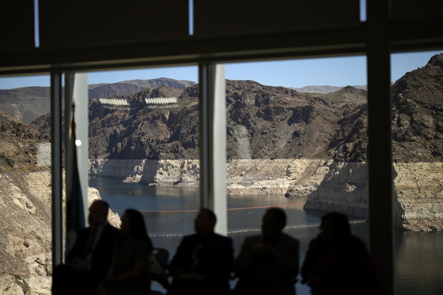 Silhouettes of people seated at a meeting with Lake Mead and the Hoover Dam visible in the background through windows. 