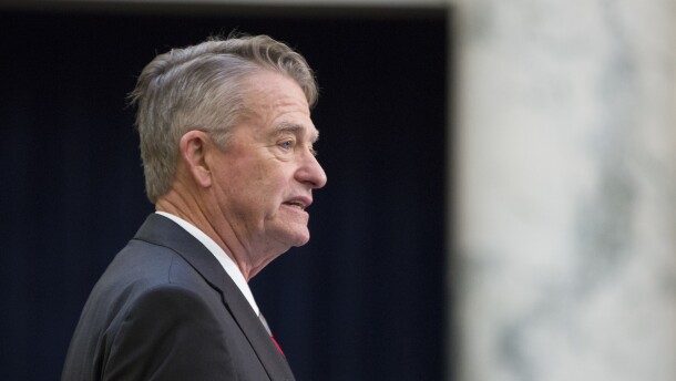 Side profile of Gov. Brad Little standing with a black suit jacket. The background is blurred. 