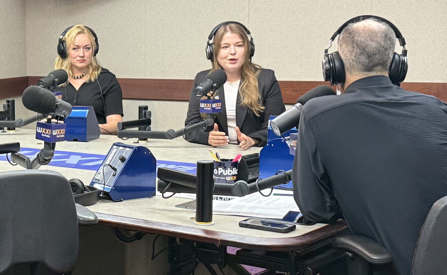 Three people wearing headphones sit at a table in a radio talk studio: a woman at left has long blonde hair and is wearing a short-sleeved black shirt and beaded necklace; a woman at center has long brown hair and is wearing a navy blazer over a white top; a man with very short dark hair at right is sitting with his back to the camera and wearing a navy shirt.