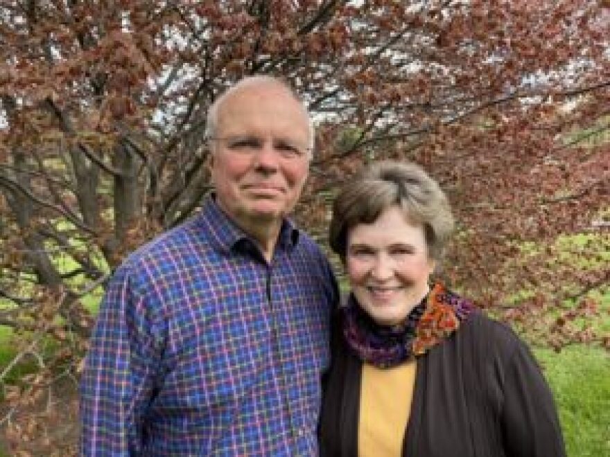 Nancy Mack stands next to her husband. They are both smiling for the camera. They stand in front of a tree with orange leaves.