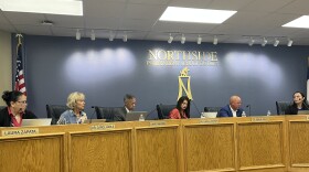 A group of women and men sit at a wooden dais with microphones. A sign behind them says Northside Independent School District.
