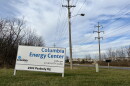 A white sign with blue lettering reads "Columbia Energy Center." The sign stands on green grass. A road and power lines are visible in the background. 