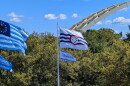 Flags blow in the wind before green trees and a blue sky. Part of a yellow bridge arch can be seen in the background.