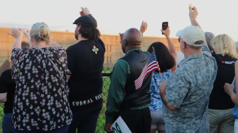 People stand with arms outstretched holding candles and signs at a fence facing a jail building