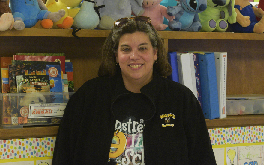 A woman with an Oakland Elementary branded jacket stands in front of a bookshelf.