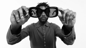 A black-and-white image of a man wearing a button-down shirt holding dark sunglasses away from his face and close to the camera.