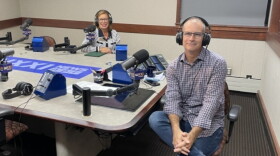 Two people wearing headphones and eyeglasses sit at a table in a radio talk studio: a woman at left has shoulder-length brown hair and is wearing a blue-and-white striped sweater with a blue collar; a balding man at right is wearing a plaid button-down shirt, blue jeans and brown shoes.