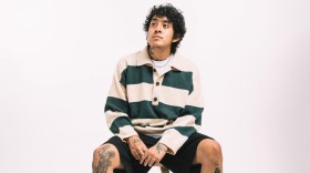 A man with tattoos on his hands and neck wearing a long-sleeved collared shirt and shorts sits on a stool against a light background.