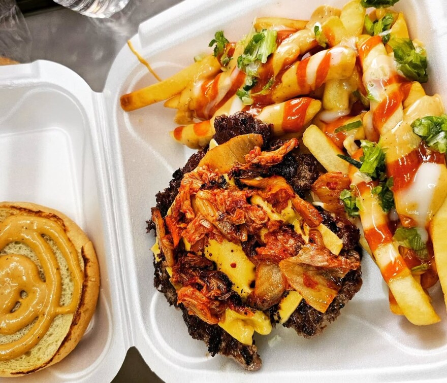 A take out container with a burger topped with vegetables and fries drizzled with sauce.