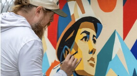 A close shot of the artist, a bearded white man, leaning towards his mural, painting the details of a face looking in the distance, and wearing a helmet. 
